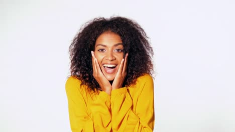 Hermosa-mujer-afro-americana-con-el-pelo-afro-en-desgaste-amarillo-sonriente,-sorprendido-agradablemente-a-la-cámara-sobre-fondo-de-pared-blanca.-Retrato-de-la-muchacha-linda-de-raza-mezclada-con-asombro