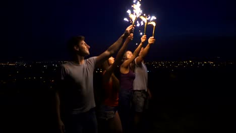 The-four-people-stand-with-firework-sticks-on-a-city-background.-night-time