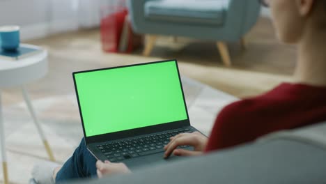 Junge-Frau-zu-Hause-arbeitet-an-einem-Laptop-Computer-mit-grünen-Mock-up-Bildschirm.-Sie-sitzt-auf-einer-Couch-in-seinem-gemütlichen-Wohnzimmer.-Über-die-Schulter-Kamera-Aufnahme