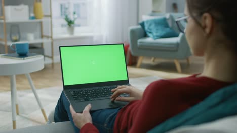 Junge-Frau-zu-Hause-arbeitet-an-einem-Laptop-Computer-mit-grünen-Mock-up-Bildschirm.-Sie-sitzt-auf-einer-Couch-in-seinem-gemütlichen-Wohnzimmer.-Über-die-Schulter-Kamera-Aufnahme