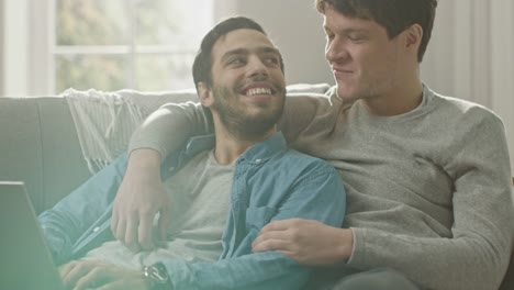 Sweet-Male-Queer-Couple-Spend-Time-at-Home.-They-are-Lying-Down-on-a-Sofa-and-Use-the-Laptop.-They-Browse-Online.-Partner's-Hand-is-Around-His-Lover.-They-Smile-and-Laugh.-Room-Has-Modern-Interior.