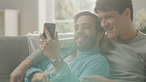 Cute-Male-Gay-Couple-Spend-Time-at-Home.-They-are-Lying-Down-on-a-Sofa-and-Use-a-Smartphone.-They-Browse-Online.-Partner's-Hand-is-Around-His-Lover.-They-Smile-and-Laugh.-Room-Has-Modern-Interior.