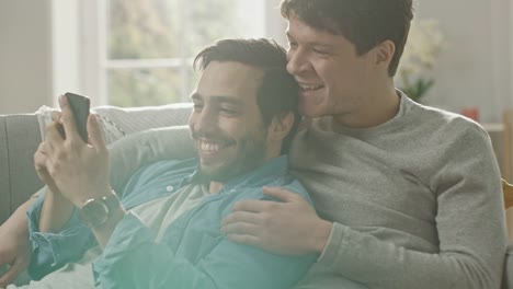 Cute-Male-Gay-Couple-Spend-Time-at-Home.-They-are-Lying-Down-on-a-Sofa-and-Use-a-Smartphone.-They-Browse-Online.-Partner's-Hand-is-Around-His-Lover.-They-Smile-and-Laugh.-Room-Has-Modern-Interior.