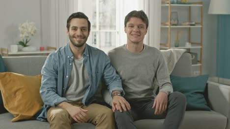 Cute-Attractive-Male-Gay-Couple-Sit-Together-on-a-Sofa-at-Home.-Boyfriend-Puts-His-Hand-on-Fiance's.-They-are-Happy-and-Smiling.-They-are-Casually-Dressed-and-Their-Room-Has-Modern-Interior.