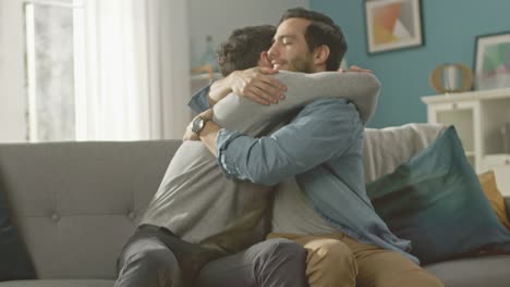 Cute-Attractive-Male-Gay-Couple-Sit-Together-on-a-Sofa-at-Home.-Boyfriends-are-Hugging-and-Embracing-Each-Other.-They-are-Happy-and-Smiling.-They-are-Casually-Dressed-and-Room-Has-Modern-Interior.