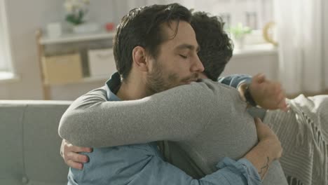 Pareja-Gay-masculino-atractivo-lindo-sentarse-juntos-en-un-sofá-en-casa.-Novios-son-Hugging-y-abrazándose-unos-a-otros.-Ellos-son-feliz-y-sonriente.-Son-vestidos-casualmente-y-sala-tiene-un-Interior-moderno.