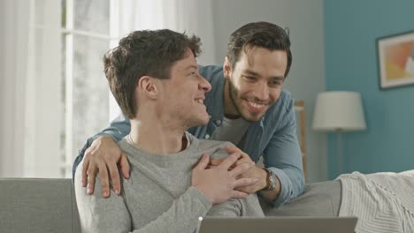 Süße-männliche-Gay-paar-verbringen-Zeit-zu-Hause.-Junger-Mann-arbeitet-auf-einem-Laptop,-seine-Partnerin-von-hinten-kommt-und-sanft-umarmt-ihn.-Sie-lachen-und-Hände-berühren.-Zimmer-ist-Modern-eingerichtet.