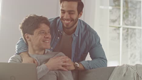 Sweet-Male-Gay-Couple-Spend-Time-at-Home.-Young-Man-Works-on-a-Laptop,-His-Partner-Comes-From-Behind-and-Gently-Embraces-Him.-They-Laugh-and-Touch-Hands.-Room-Has-Modern-Interior.