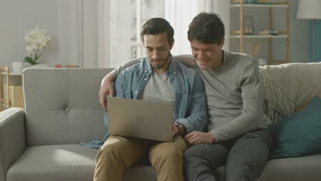 Entzückende-männliche-Gay-paar-verbringen-Zeit-zu-Hause.-Sie-sitzen-auf-einem-Sofa-und-das-Notebook-nutzen.-Stöbern-sie-Online.-Partner-legt-seine-Hand-um-seine-Geliebte.-Zimmer-ist-Modern-eingerichtet.