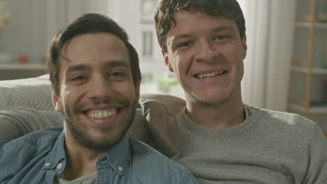 Portrait-of-a-Cute-Male-Queer-Couple-at-Home.-They-Sit-on-a-Sofa-and-Look-at-the-Camera.-Partner-Embraces-His-Lover-from-Behind.-They-are-Happy-and-Smiling.-Room-Has-Modern-Interior.