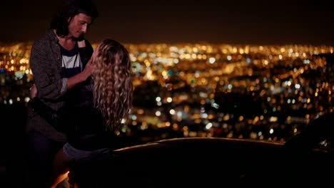 Young-couple-hugging-and-looking-at-night-city-lightsnext-to-a-convertible