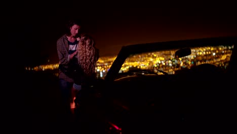 Young-couple-hugging-and-looking-at-night-city-lightsnext-to-a-convertible