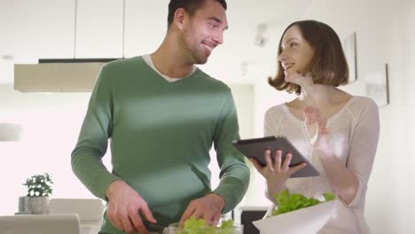 Happy-couple-is-preparing-healthy-food-in-the-kitchen-at-home-while-checking-a-tablet-computer.