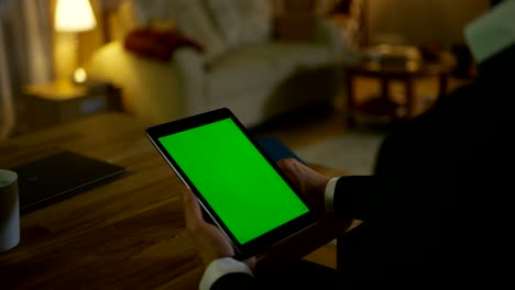 At-Home-Man-Sits-at-His-Desk-and-Holds-Tablet-Computer-with-Green-Screen-on-it.-His-Apartment-is-Done-in-Yellow-colours-and-is-Warm.