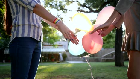 LGBT-girlfriends-stand-together-holding-hands-with-inflatable-balloons-and-bracelet-in-backlight