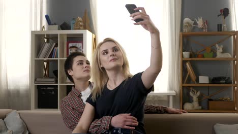 Zwei-junge,-schöne-Mädchen-sitzen-auf-der-Couch-und-machen-Selfies,-Lesben,-LGBT,-blond,-brünett-60fps