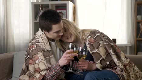 Two-young,-beautiful-girls-are-sitting-on-the-couch-wrapping-with-warm-plaid-blanket,-drinking-wine-and-hugging,-cuddle-60-fps