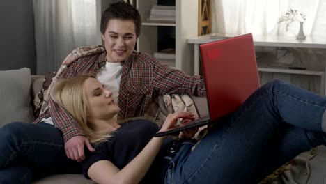 Young-blond-woman-lies-on-the-lap-of-a-brunette-and-using-red-laptop,-cosiness,-cuddles,-plaid-60-fps