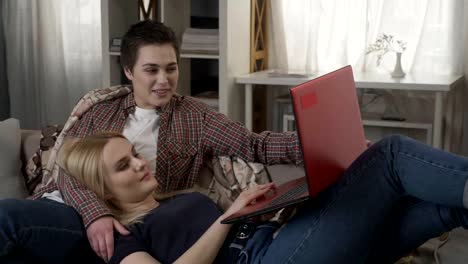 Young-blond-woman-lies-on-the-lap-of-a-brunette-and-using-red-laptop,-cosiness,-cuddles,-plaid,-laughing-60-fps