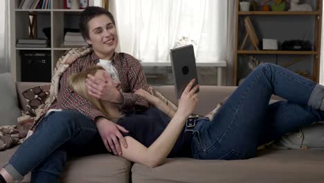 Lesbisches-Paar-ruht-sich-auf-der-Couch,-mit-Tablet-PC,-Mädchen-mit-kurzen-Haaren-sanft-streichelt-ihr-Partner-Gesicht-sprechen,-Familienidylle,-LGBT-60-fps