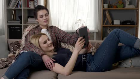 Lesbian-couple-is-resting-on-the-couch,-using-tablet-computer,-scrolling-photos-on-tablet,-holding-hands,-cuddling-60-fps