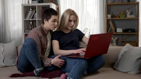 Two-young-lesbian-girls-are-sitting-on-the-couch,-using-a-computer,-scrolling-the-internet,-shopping-online,-the-blonde-maliciously-closes-the-laptop,-quarrel-60-fps