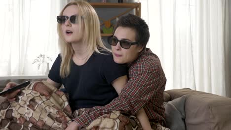 Zwei-junge-lesbische-Mädchen-sitzen-auf-der-Couch,-in-eine-warme-Decke-bedeckt-vor-der-3d-Film-über-die-Fernbedienung,-schockiert,-überrascht-60fps