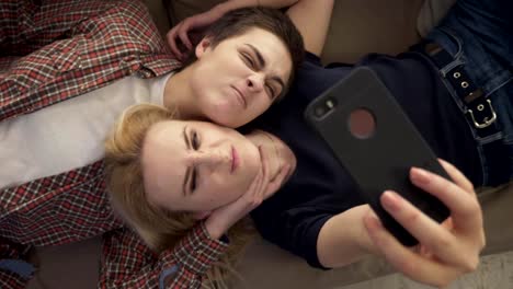 Two-young-girls-lie-on-the-couch,-do-selfie-on-a-smartphone,-grimace,-make-funny-faces,-top-shot-60-fps