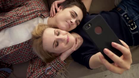 Two-young-girls-lie-on-the-couch,-do-selfie-on-a-smartphone,-smiling,-lovers,-lgbt,-young-couple,-top-shot-60-fps