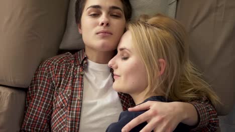 Two-young-lesbian-girls-lie-on-the-couch,-hug,-cuddle,-sleep,-girl-with-short-hair-looks-at-the-camera,-lgbt-family-concept,-top-shot-60-fps
