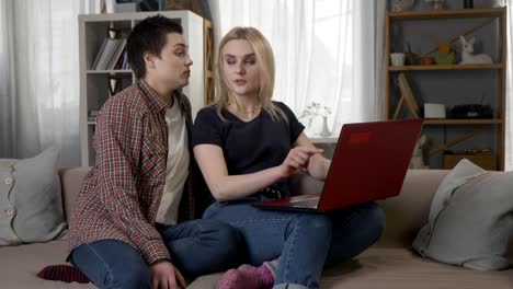 Two-young-lesbian-girls-are-sitting-on-the-couch,-using-a-computer,-scrolling-the-internet,-shopping-online,-smiling,-talking