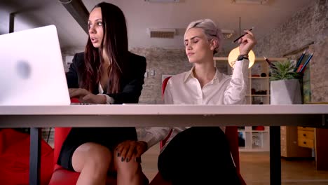 Young-confident-blonde-woman-touching-knee-of-other-woman-in-modern-office,-lesbian-concept,-sitting-at-table-with-laptop