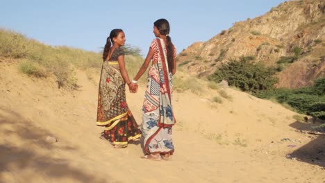 Señoras-mano-andando-hablando-desierto-Rajasthani-tradicional-vestido-de-verano-piedra-sol-arena-mano-vinculación-amor-comparte-espalda-ancha-tiro-medio-siguen-estabilizado-cardán-colina-sari-Vestido