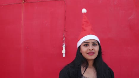 Indian-woman-with-Santa's-hat-taking-selfies-photo-with--mobile-cell-phone,-cheerful-and-excited,-with-red-background