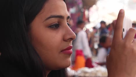 Close-up-of-a-pretty-woman-traveller-using-her-smart-phone-mobile-device-during-her-vacation-in-Rajasthan-Pushkar-handheld-side-profile-smile-content-happy-joy-touch-screen-street-crowded-out-of-focus