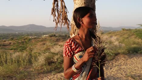 Beautiful-Indian-lady-under-a-tree-waits-for-lover-contemplating-worried-thinks-broods-with-fern-in-hand-fidgety-nervous-clandestine-meeting-point-date-outdoor-summer-hot-sun-sunlight-nature-handheld