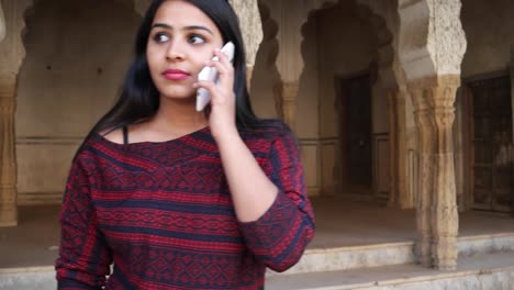 Handheld-shot-of-young-woman-on-her-mobile-phone-cellphone-smart-communicate-talks-touch-screen-talks-message-texts-share-network-smiles-in-front-of-Indian-architecture-column-arch-backdrop-close-up