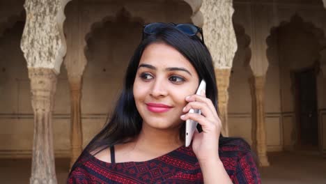 Handheld-shot-of-young-woman-on-her-mobile-phone-cellphone-smart-communicate-talks-touch-screen-talks-message-texts-share-network-smiles-in-front-of-Indian-architecture-column-arch-backdrop-close-up