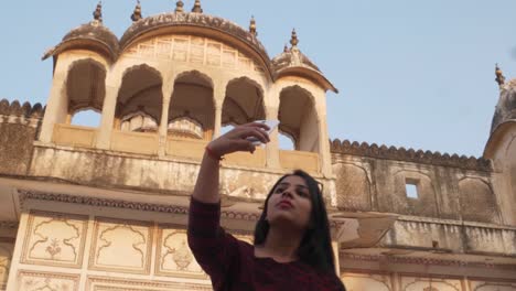 Handheld-shot-of-tourist-woman-in-front-of-Indian-Hindu-temple-taking-selfie--photo-video-on-her-mobile-smart-phone-happy-share-facade-architecture-ancient-holy-religious-fort-castle-palace-Rajasthani