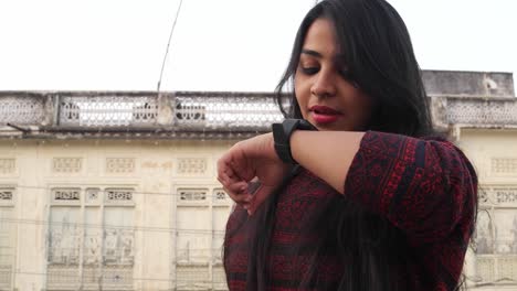 Young-attractive-woman-busy-with-smart-watch-talks-listens-communicates-smiles-laughs-happy-joy-looks-at-the-camera-with-a-traditional-local-Rajasthani-house-facade-in-the-backdrop-handheld-mid-shot