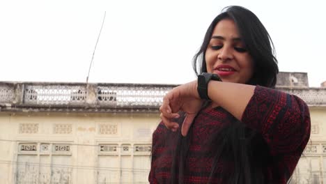 Young-attractive-woman-busy-with-smart-watch-talks-listens-communicates-smiles-laughs-happy-joy-looks-at-the-camera-with-a-traditional-local-Rajasthani-house-facade-in-the-backdrop-handheld-mid-shot