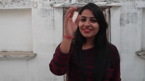 Young-woman-enters-and-exits-an-old-building-doorway-with-folded-joined-hands-to-namaste,-a-traditional-Indian-greeting-for-respect-laughs-smiles-points-hands-youngster-waves-jovial-elders-handheld