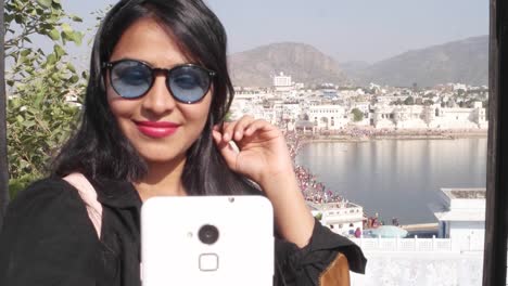 Handheld-Frau-nimmt-Selfie-Foto-Video-mit-ihrem-mobilen-Handy-Gerät-von-einem-Panorama-Aussichtspunkt-Blick-des-Heiligen-Pushkar-See,-eine-heilige-Stadt-für-Hindu-Pilger-Mela-Festival-Baden-Ghats-Tempel