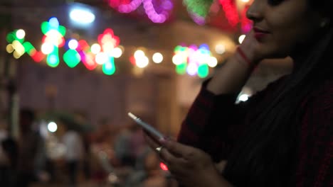 Young-Indian-woman-on-a-touch-screen-smart-mobile-phone-texts-messages-types-shares-photo-video-calls-in-front-of-a-festival-colorful-bright-lights-out-of-focus-in-the-background-celebration-mela-love