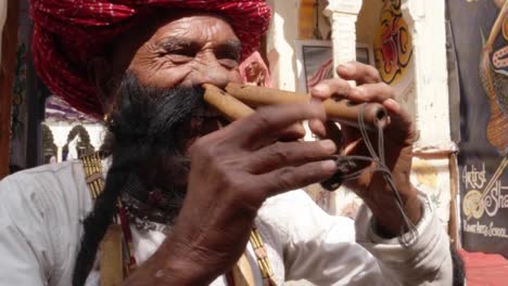 Hand-held-CU-Rajasthani-elderly-male-plays-the-flute-with-his-nose-in-front-of-a-painted-temple-archway,-with-big-moustache-wearing-traditional-attire