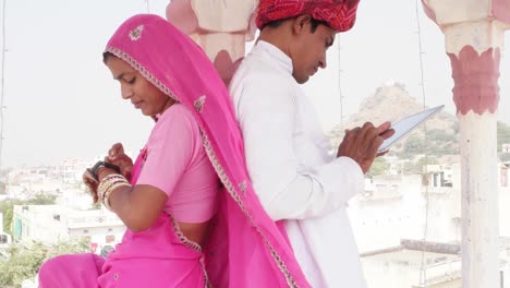 Couple-busy-on-their-smart-watch-and-tablet-sepreately-ignoring-with-their-backs-to-each-other-under-a-chattri-canopy-in-Rajasthan-India