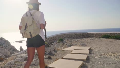 Portrait-of-a-beautiful-young-tourist-(girl),-looks-at-an-lighthouse,-with-a-backpack,-in-a-straw-hat,-a-white-house-backdrop.