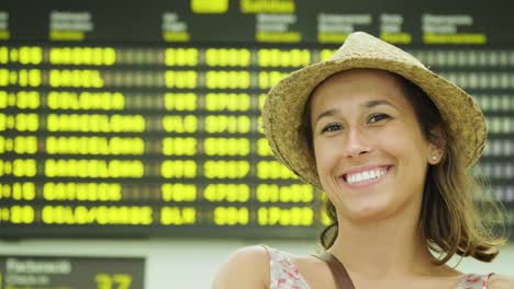 Schönes-junges-Mädchen-am-Flughafen,-mit-Blick-auf-den-Flugplan-mit-Stroh-Hut,-in-einem-Kleid,-lächelnd.