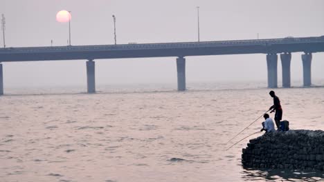 Fisherman-fishing-in-the-sea-against-the-modern-bridge-during-evening