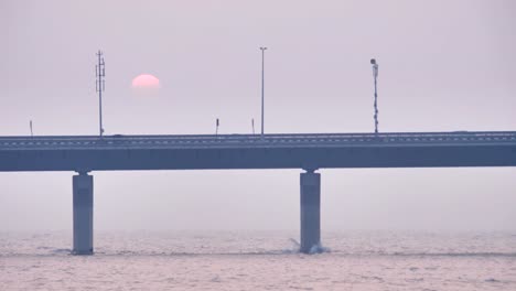 Fahrzeuge,-die-auf-einer-modernen-Brücke-vorbei,-während-im-Hintergrund-Sonne-untergeht